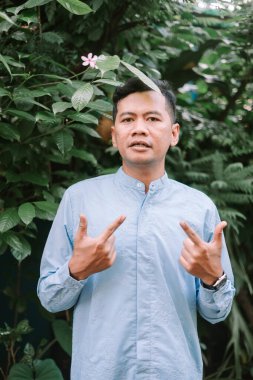 Dost canlısı bir adamın portresi açık hava ortamında, elleriyle el kol hareketi yapıyor. O, şık bir mavi gömlek giyen verimli tropikal yeşillik ile çevrilidir. Doğal ortamda sakin, neşeli ve olumlu bir ifade yakalamak..