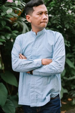 Düşünceli görünümlü Asyalı bir adam dışarıda duruyor, kollarını kavuşturmuş, sakin ve düşünceli ifadesini sergiliyor. Şık, açık mavi bir gömlek ve narin bir nakış giyerek kendine güven ve zarafet saçar. Doğal yeşillik arka planı ekle