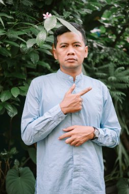 Açık mavi gömlekli Asyalı bir adam dışarıda duruyor, yan tarafı işaret ediyor. Bereketli yeşilliklerle çevrili, dingin ifadesi ve odaklanmış bakışları büyüleyici bir portre oluşturuyor. Rehberlik, sunum ya da doğal sağlık kavramlarını açıklamak için mükemmel.