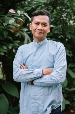 Gülümseyen Asyalı bir adam dışarıda kollarını kavuşturmuş bekliyor. Geleneksel açık mavi bir gömlek giyiyor. Arkaplanda, sakin ve doğal bir ortam yaratan yemyeşil bitkiler yer alıyor. İfadesi güven ve olumlu bir görünüm yayıyor.. 