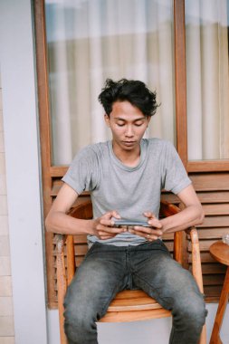 Genç adam bir telefon oyununa dalmış, verandadaki bir sandalyede mobil cihazıyla dinleniyor.