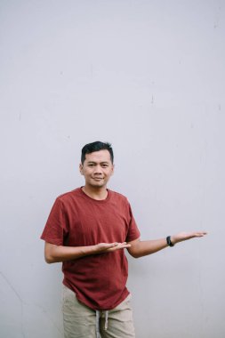 Asyalı bir adam avuç içi açık bir şey sunuyor. Günlük giysi, gri beton arka plan, tarafsız ifade ve davetkar jest..