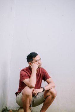 Düşünceli Asyalı bir adam, eli yüzü üzerinde, derin bir düşüncelere dalmış, bir duvara doğru çömeliyor.