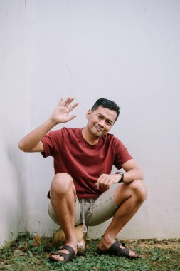 Sıradan bir Asyalı adam çömelir ve selam verir. Minimum, tarafsız bir arka plana sıcak bir şekilde gülümser. Arkadaşlık ve cana yakın cazibe..