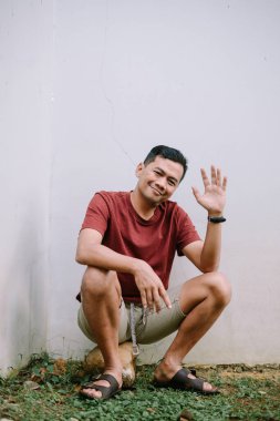 Dost canlısı Asyalı bir adam beyaz bir duvarın yanında oturuyor ve el sallıyor. Neşeli ve misafirperver görünüyor..