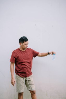 Çöpe atılmış plastik bir şişeyi düz bir duvara karşı incelikle inceleyen bir adam. Odaklanma kirlilik ve ekolojik sorumluluktur.