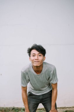 Gri gömlekli arkadaş canlısı genç bir adam, tarafsız bir duvara karşı mutlu ve neşeli bir ifadeyle öne eğiliyor..