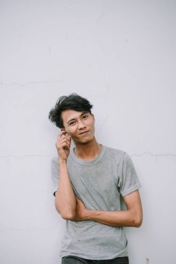 Düşünceli genç bir adam, günlük giysiler içinde, beyaz bir duvara yaslanmış, rahatlamış bir güven ve sessiz bir tefekkür hissi yayıyor.