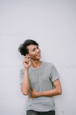 Gülümseyen genç adam duvarda gri gömlekli ve mutlu bir ifadeyle poz veriyor.