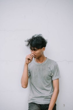 Beyaz bir duvara yaslanmış gri tişörtlü dalgın genç bir adam. Minimalist portre modern ve düşünceli bir ruh hali sergiliyor..