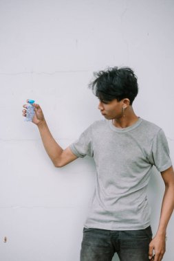 Genç bir adam geri dönüşümlü bir su şişesi tutuyor ve basit dokulu bir duvara karşı düşünceli görünüyor. Sürdürülebilirlik ve susuzluğa odaklan.