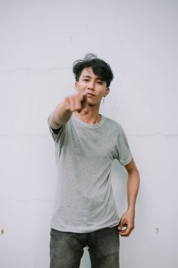 Modayı takip eden genç bir adam, minimalist beyaz bir duvara karşı anlamlı bir hareketle kamerayı işaret ediyor. Gündelik ve modern stil.