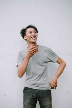 Gri gömlekli mutlu bir genç adam neşeli bir ifadeyle dokulu beyaz bir duvara gülümsüyor.