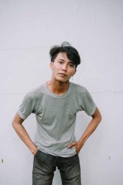 Düşünceli Asyalı bir adam elleri cebinde yıpranmış beyaz bir duvarın yanında poz veriyor..