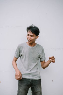 Gri tişörtlü, beyaz arka planda dikilen dalgın Asyalı genç adam.