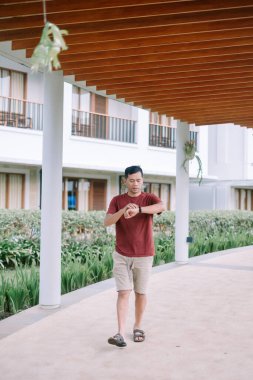 Modern bir tropikal tatil köyünde yürüyüş yolunda yürürken akıllı saatini kontrol eden bir adam. Rahat ve rahat bir şekilde giyinmiş, tatilin tadını çıkarıyor..