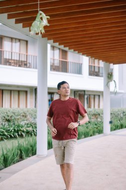 Asyalı adam otelin modern koridorunda düşünceli bir şekilde yürüyor, tatiline doğru ilerliyor.