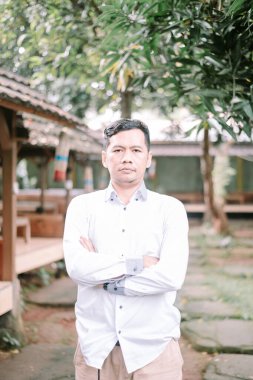 Kendine güvenen Asyalı adam kollarını kavuşturmuş dışarıda duruyor, azim ve başarı gösteriyor. Doğal ortamdaki iş portresi.