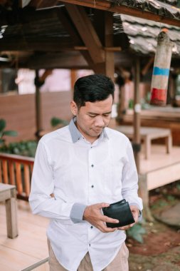 Asyalı bir adam açık havada cüzdanını incelikle inceliyor, muhtemelen mali kaygılarını yansıtıyor ya da mevcut bütçesini değerlendiriyor.