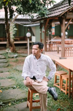 Asyalı gömlekli bir adam açık havada bir sandalyede oturuyor, el ele tutuşmuş, bir an düşüncelere dalmış.