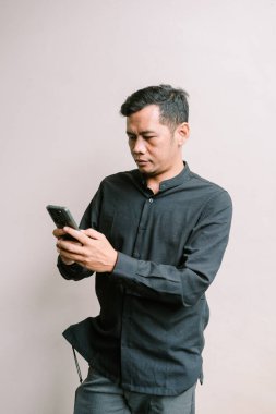 Siyah tişörtlü Asyalı adam akıllı telefonunu ciddi bir ifadeyle dikkatle kullanıyor. Tarafsız arkaplanda izole.