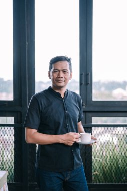 Olgun bir Asyalı adam evdeki pencerenin yanında bir fincan kahvenin tadını çıkarıyor. Günlük kıyafetlerle, iç mekanlarda rahatlamak.