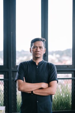 Ciddi bir ifade takınan bir adam, kollarını pencereye bağlamış, kendinden emin bir şekilde durur.. 