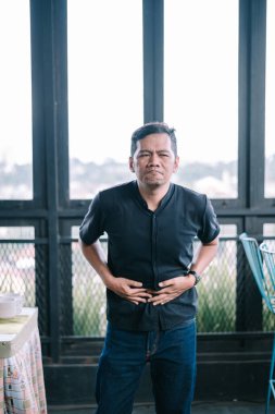 Asyalı bir adam acı içinde karnını tutar ve rahatsızlık ifadesi gösterir. Mide ağrısı ya da sindirim sorunu olabilir..