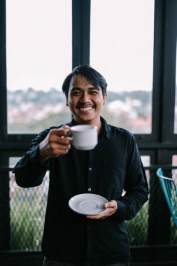Neşeli bir adam elinde bir fincan kahveyle balkonda samimi bir gülümseme ve şehir manzaralı sıcak bir içecek sunuyor..