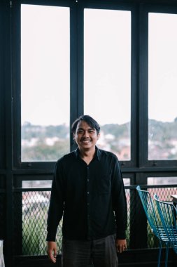 Gülümseyen bir adam pencerenin yanında durur, arka planda hafifçe görünen bir şehir manzarasından memnuniyet yayar..