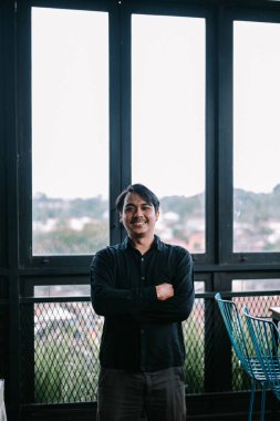 Çapraz kollu genç adam geniş pencereli modern bir ortamda güvenle gülümsüyor.