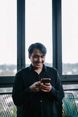 Pencerenin yanında akıllı telefon kullanan mutlu yüzlü bir adam. Bağlı ve gülümsüyor.