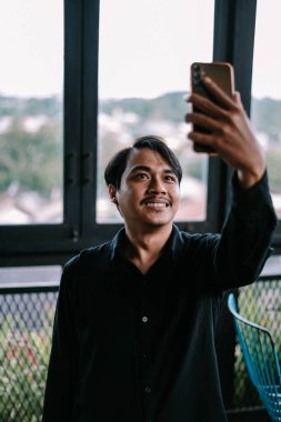 Pencerenin yanında akıllı telefon kullanarak selfie çeken mutlu Asyalı portre.