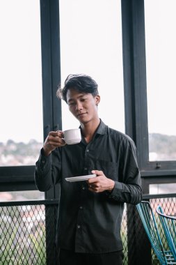 Genç Asyalı bir adam bir fincan kahvenin tadını çıkarıyor. Sakin ve sakin bir ortamda düşünceli bir şekilde pencerenin yanında duruyor.