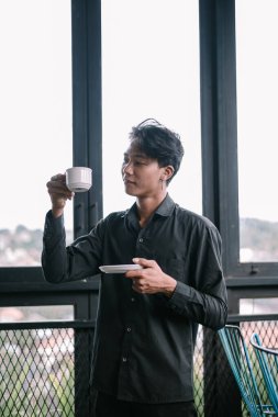 Genç Asyalı adam evinin penceresinde kahve içerken sessiz bir anın tadını çıkarıyor. Onun sakin tavrı bir huzur hissi uyandırıyor.