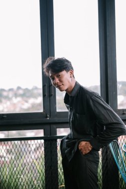 Genç bir adam pencereden dalgın dalgın bakıyor. Karanlık bir kıyafet giyiyor, modern bir tarz yayıyor ve düşünceli bir duruş sergiliyor..