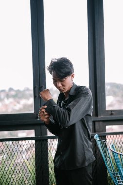 Siyah gömlekli genç Asyalı bir adam pencerenin yanında giyiniyor, güne hazırlanıyor. Kelepçelerini düşünceli bir ifadeyle ilikliyor..