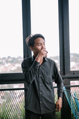 Sıradan gömlekli düşünceli genç Asyalı adam şehir manzaralı pencereden dışarı bakıyor. Huzurlu ve düşünceli ifade.