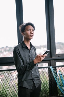 Genç bir adam cep telefonunu evin içinde tutarken, düşünceli bir şekilde pencerenin yanında, düşüncelere dalmış..