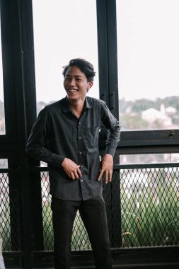 Koyu renk gömlekli neşeli Asyalı bir adam korkuluklara yaslanıyor ve neşeyle gülümsüyor.