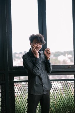 Genç adam şehir manzaralı pencerenin yanında poz veriyor. Şaşırmış ve mutlu görünüyor..