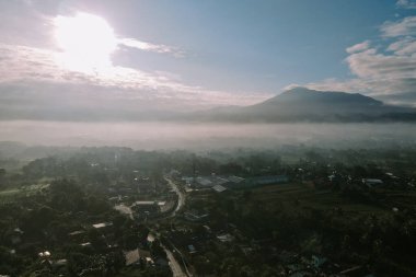 Dağların arasına yerleşmiş, sabah güneşiyle yıkanmış sisli bir köyün huzurlu manzarasını gösteren bir hava görüntüsü..