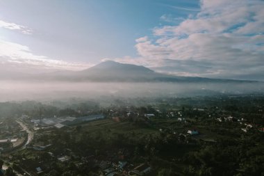 Huzurlu bulutlu bir sabahta sisli bir dağ sırasının altında yuva yapmış bir köyü gösteren nefes kesici hava perspektifi. Sahne, doğanın sükunetini yakalar..
