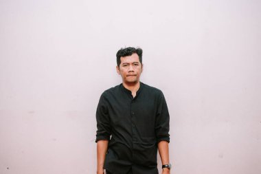 Koyu renk gömlekli düşünceli bir adam tarafsız bir duvara poz veriyor. Görüntü onun ciddi ifadesini ve şık görünümünü yakalıyor.