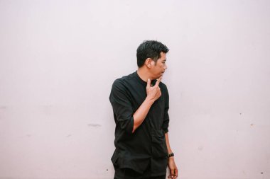 Siyah tişörtlü düşünceli Asyalı bir adam temiz beyaz bir duvara karşı düşünceli bir şekilde bakar ve fotokopi alanı bırakır.