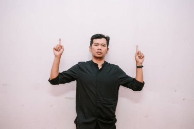 Siyah tişörtlü Asyalı bir adam düz bir duvara doğru boşluğu işaret ediyor. Reklamlar, modeller ve promosyonlar için harika.