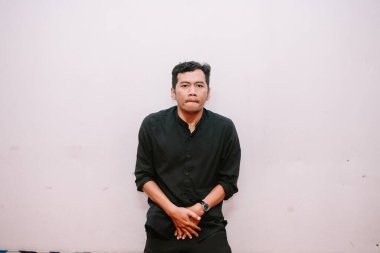 Siyah gömlekli düşünceli Asyalı bir adam düz bir duvara karşı dalgın dalgın duruyor. Stüdyo çekimi.