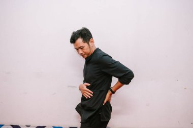 Sırt ağrısından muzdarip bir adam, düz duvara karşı duruyor. Sıradan kıyafetler giyiyor ve rahatsız görünüyor..