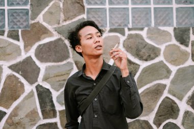 Sigara içen şık bir genç adam, taş bir duvara karşı poz verir, düşüncelere dalma ve kentsel serinlik hissi taşır..