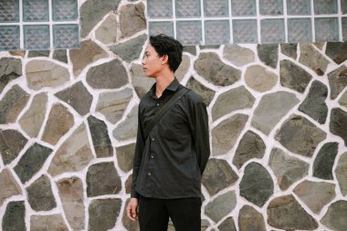 Şık giyinmiş, düşünceli genç bir adam cam aksanlı, eşsiz taş bir duvarın önünde duruyor..